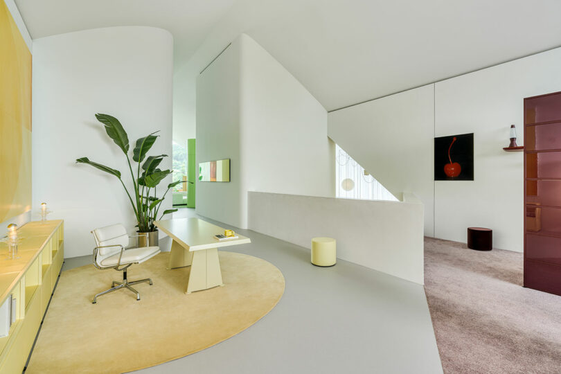 Spacious modern office interior with a white desk, ergonomic chair, potted plant, yellow cabinets, and a large circular rug, featuring minimalistic design elements and wall art.