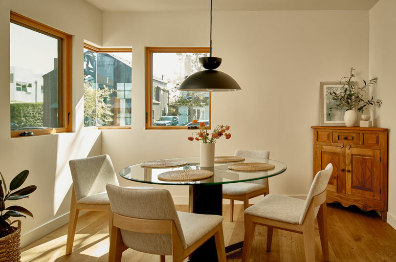 A sunlit dining area with three windows, a glass round table, four cushioned chairs, a black pendant light, a wooden sideboard with plants, and a framed picture on a white wall.