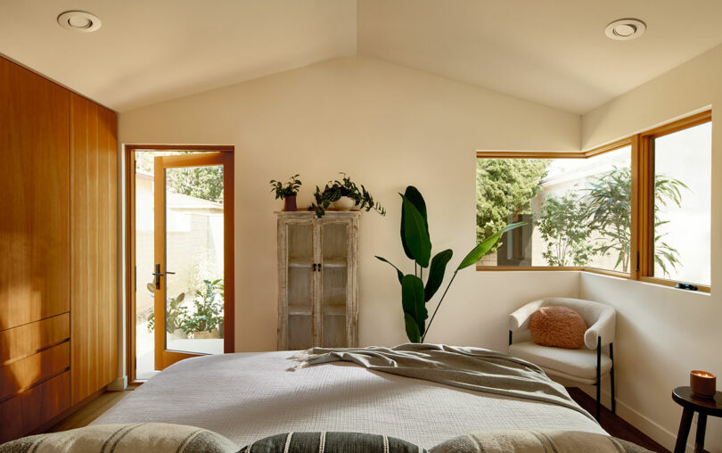 A modern bedroom with a bed, beige bedding, wooden wardrobe, potted plants, a chair with a cushion, and large windows. Sunlight illuminates the room through the windows and an open door.