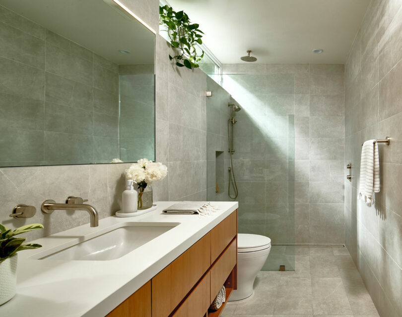 Modern bathroom with large mirror, white countertop, wooden vanity, potted plants, and walk-in shower with glass partition.
