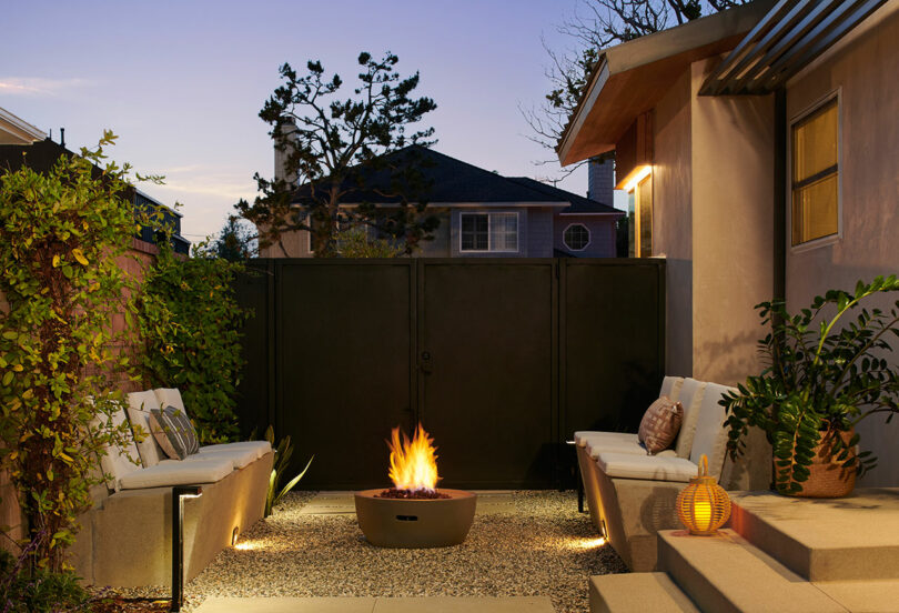A cozy outdoor patio at dusk features a central fire pit, concrete seating areas with cushions, and a lantern on the ground, surrounded by plants and privacy walls.