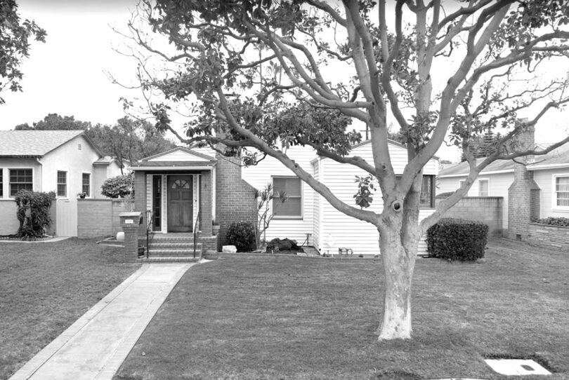 A black-and-white image of a small house with a front porch, a large tree in the yard, and a concrete pathway leading to the entrance. The house has siding and is between two similar houses.