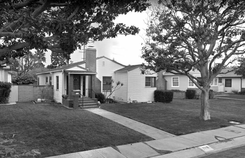A black-and-white photo of a suburban neighborhood showing a single-story house with a brick chimney, a well-maintained front lawn, and a tree in the front yard, surrounded by other similar houses.