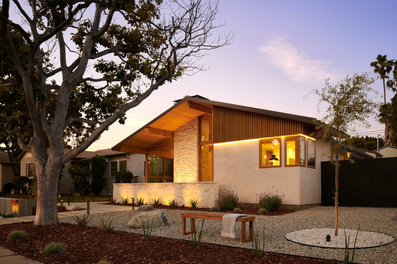 Modern one-story house with large windows, a slanted roof, and exterior lighting at sunset. A garden with rocks and a wooden bench is in the foreground, with trees and neighboring homes nearby.