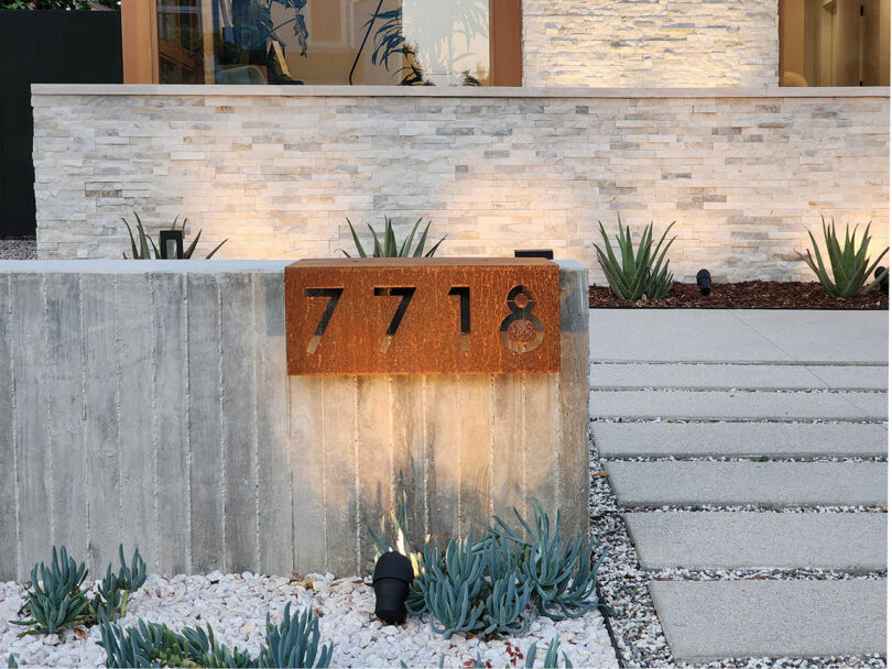 A modern house exterior with a concrete wall featuring a rusted metal house number sign "7718." The wall is surrounded by small rocks and succulents, with steps leading to the entrance.