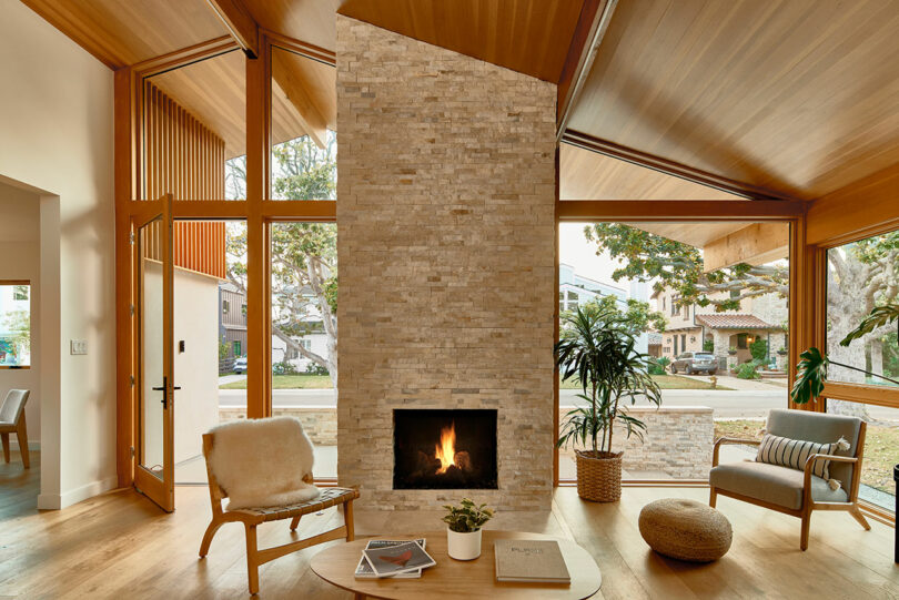 A modern living room with a central stone fireplace, wooden flooring, and large windows. Furnishings include a chair, coffee table, potted plant, and pouf. Natural light fills the space.