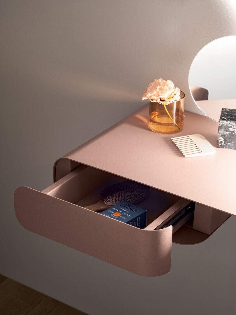 A pink metal wall-mounted desk with an open drawer reveals a hairbrush and medication. A flower in a vase, a comb, and a mirror are placed on the table's surface.