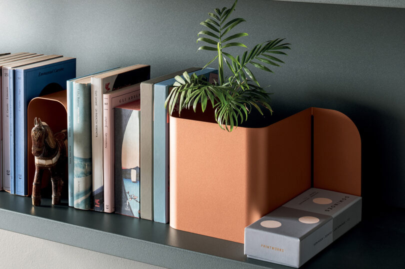 A bookshelf with books, a potted plant, a metal orange bookend, a small horse figurine, and two rectangular boxes arranged neatly on a grey shelf.