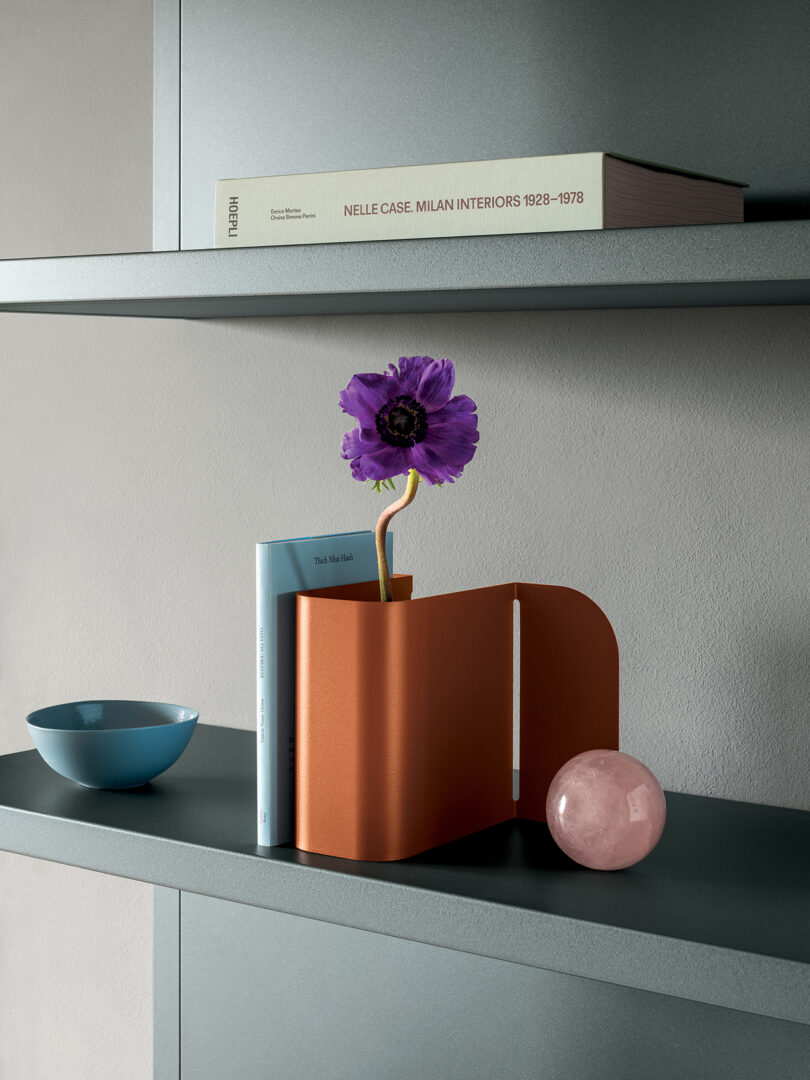 A bookshelf with an orange metal bookend, holding two books and a purple flower. Nearby are a blue bowl, a pink sphere, and a larger book titled "NELLE CASE MILAN INTERIORS 1928–1978.