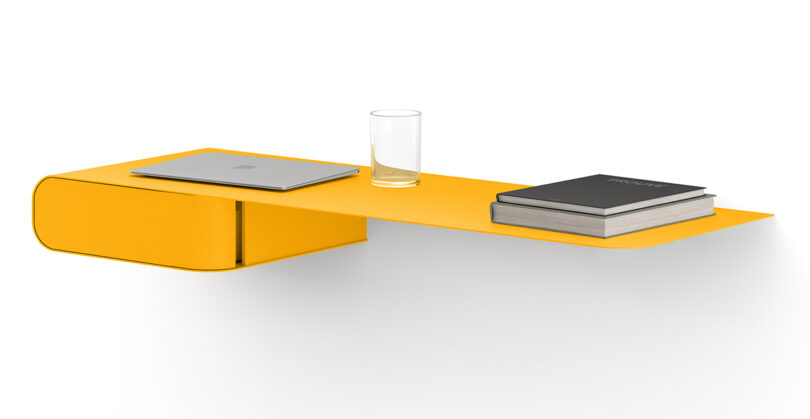 A minimalist yellow metal wall-mounted desk holds a closed laptop, a glass of water, and two books.