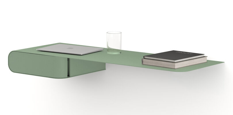 A minimalist green metal wall-mounted desk holds a closed laptop, a glass of water, and two books.