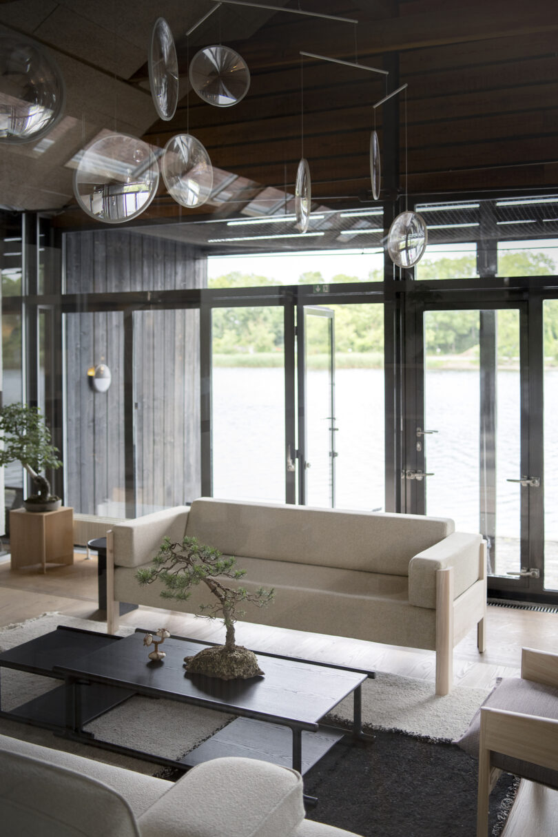 A modern living room with minimalist decor, featuring beige sofas, a bonsai tree on a black coffee table, large windows overlooking water, and hanging glass ornaments