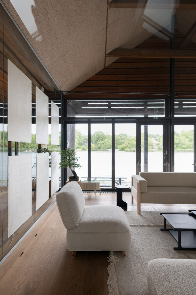 Modern living room with large windows overlooking a water view. Neutral-colored furniture, artwork on the walls, and a minimalist decor style. Light wood flooring and an area rug are present