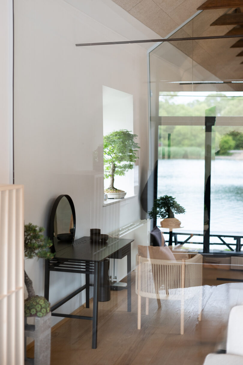 Modern interior with glass walls, featuring minimalistic furniture, two bonsai trees, and a view of a lake outside