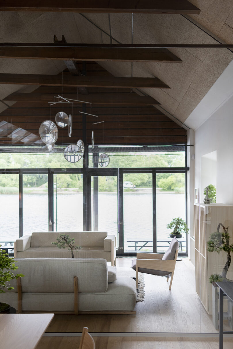 A modern living room with wooden beams, large windows overlooking a river, beige sofas, a wooden chair, and indoor plants