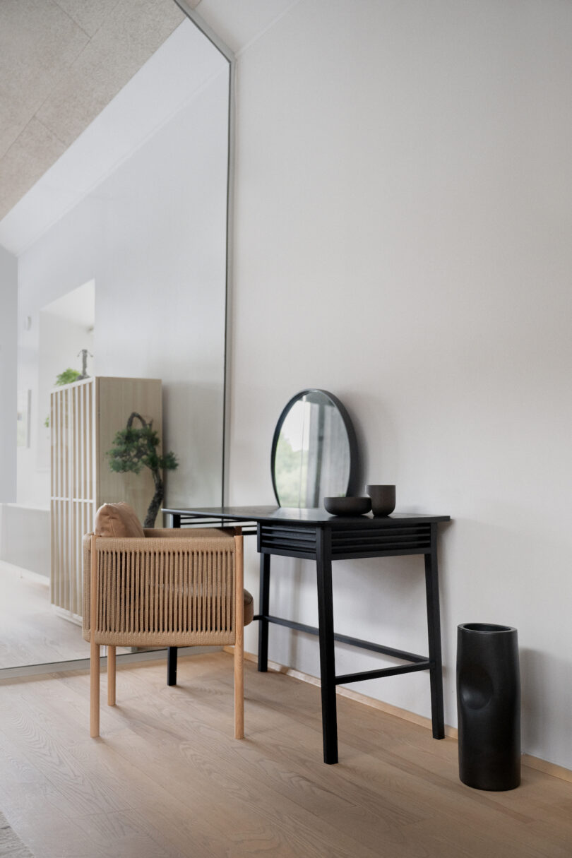A minimalist room features a wooden chair, black writing desk with a mirror, a small vase with greenery, and a large mirror along one wall. Nearby are a floor-standing black vase and a shelving unit
