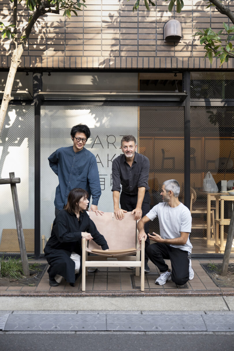 Four people gather around a wooden chair outside a building with large windows. Three are standing, one is kneeling, all appear to be discussing the chair. Urban setting with trees in the background