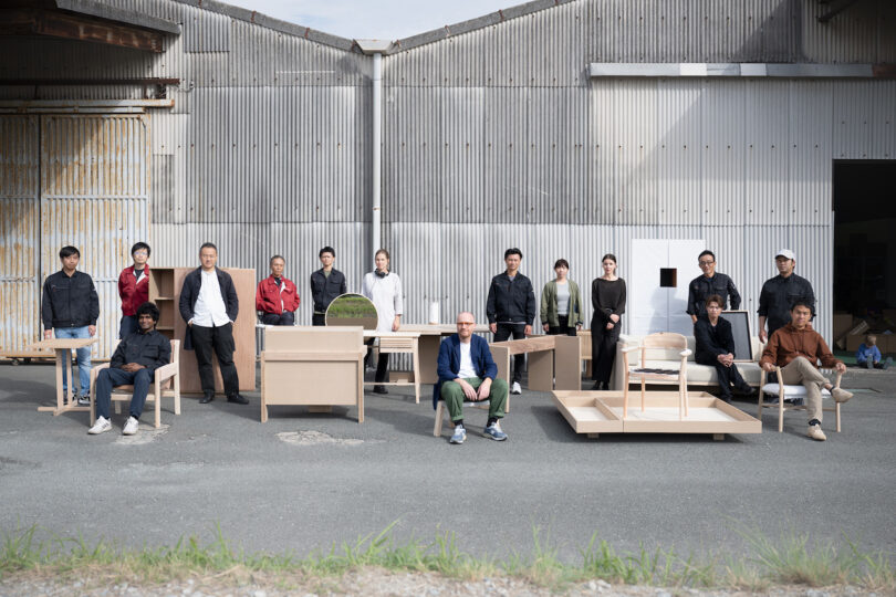 Group of people posing with various wooden furniture pieces in front of a corrugated metal warehouse
