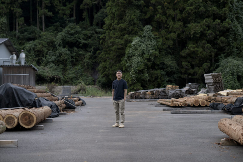 A person stands on a paved area surrounded by stacked logs and lumber, with dense foliage in the background