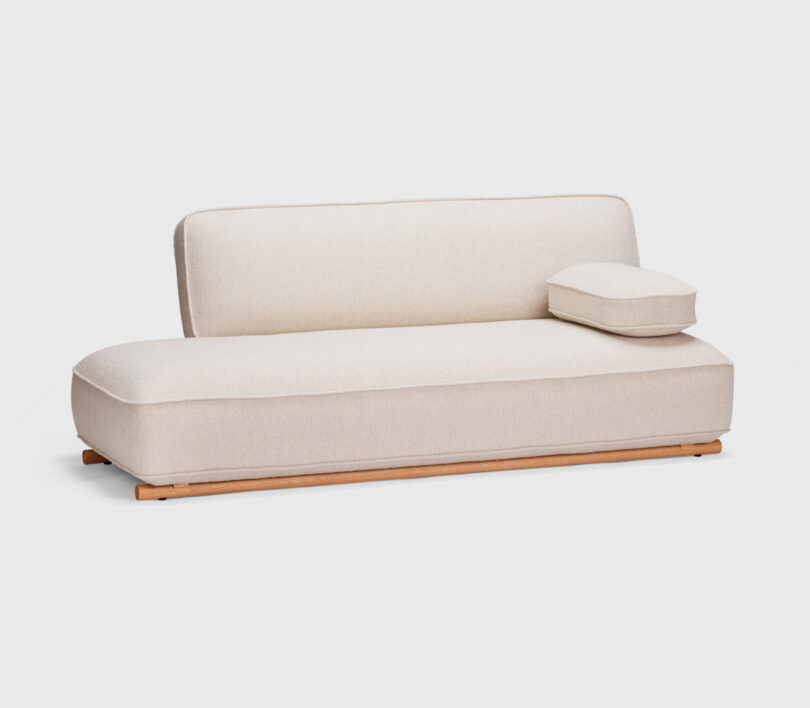 A minimalist, light beige sofa with a boxy design and a single armrest cushion, supported by a wooden base