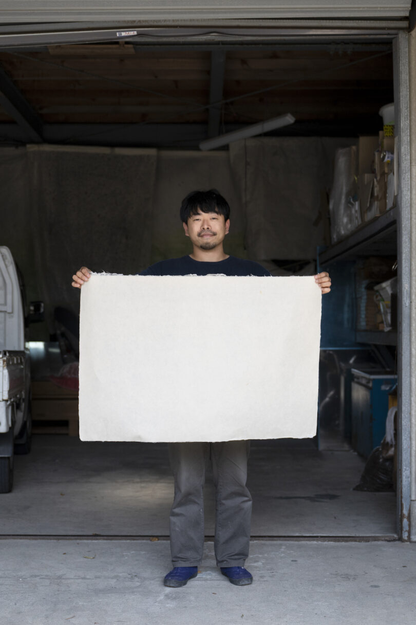 A person stands in a garage, holding up a large sheet of blank paper in front of them, with both hands extended outward, displaying it fully.