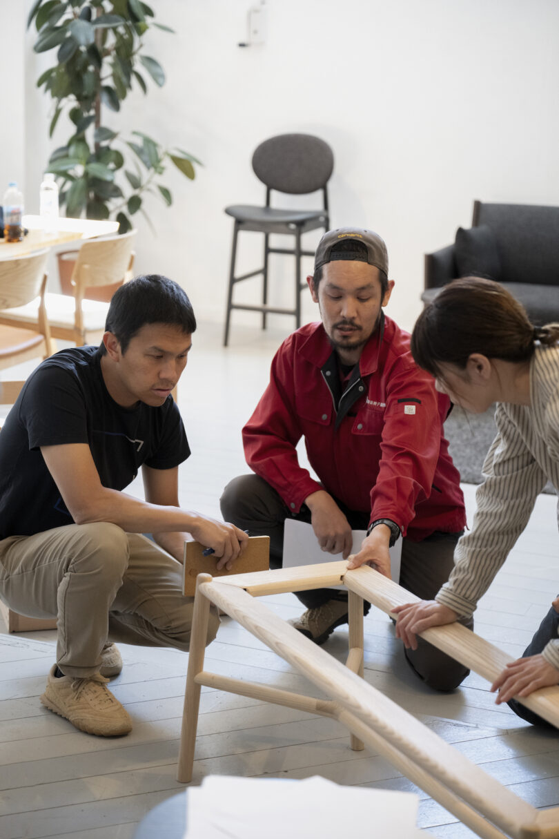 Three individuals assemble a wooden furniture piece in a room with a plant, chair, and table in the background