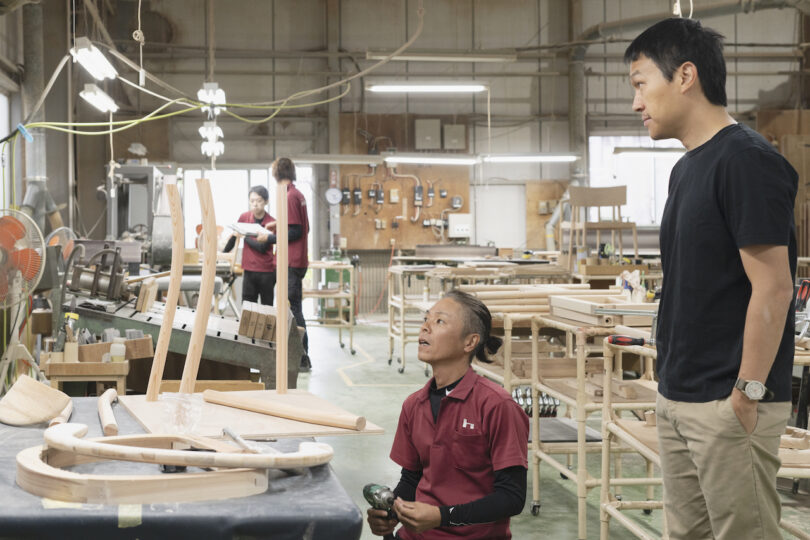 Two people in a workshop are observing wooden furniture pieces. A man in a red shirt is kneeling, holding a tool, while another man in a black shirt stands nearby. In the background, others are working
