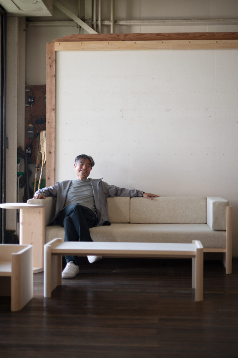 A person is sitting on a beige sofa in a minimalistic room with wooden furniture, including a coffee table and a small side table. The room has a light-colored wall and a wooden floor