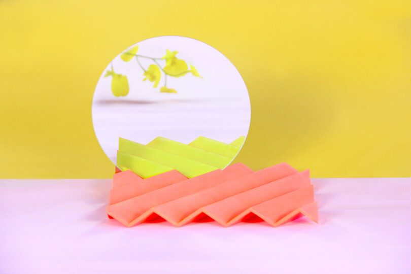 a tabletop mirror with a zigzag base on a pink surface with a yellow background behind