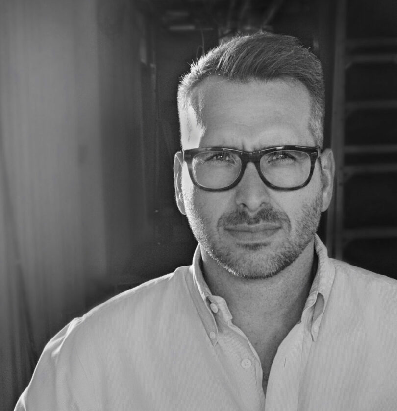 A man with short hair and glasses is wearing a button-up shirt. He looks directly at the camera with a neutral expression. The background is dimly lit and out of focus