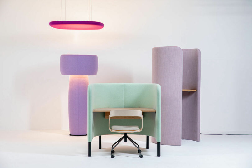 A minimalistic workspace featuring a cushioned green desk cubicle with a wooden chair, a tall purple lamp, and a standing partition with a built-in desk, all against a white background