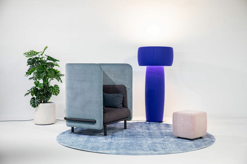 A modern seating area with a gray enclosed chair, a purple floor lamp, a potted plant, and a small pink ottoman, all arranged on a blue circular rug against a white background