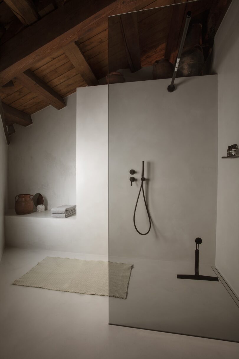 A minimalist bathroom featuring a glass shower, white walls, and wooden ceiling beams