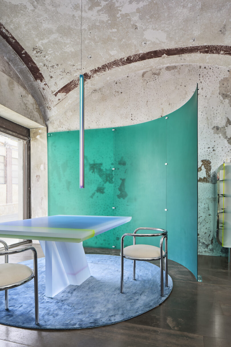 A modern dining area with a minimalist table, two chairs, a blue rug, and a green curved partition. An overhead pendant light hangs from a textured, arched ceiling