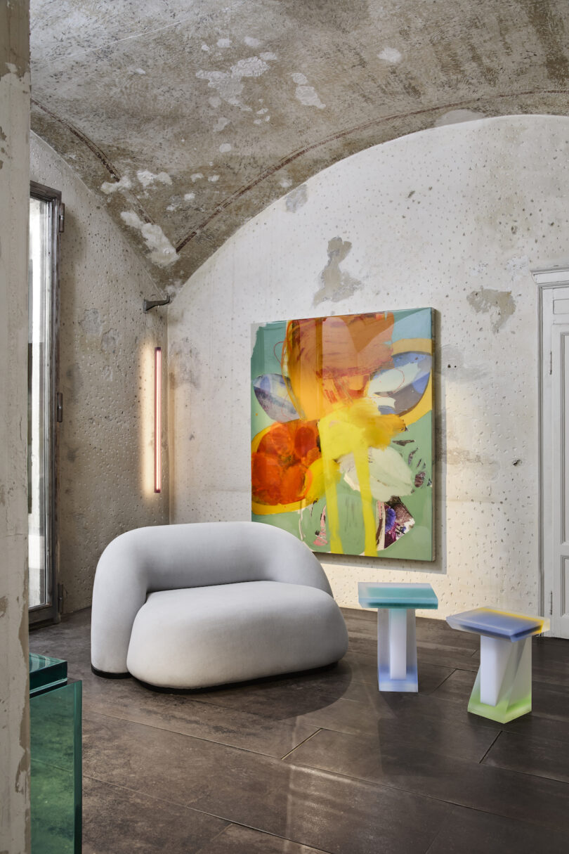 A modern room with a plush, grey, rounded chair, two colorful side tables, abstract painting on the wall, and vertical LED light on a textured concrete wall