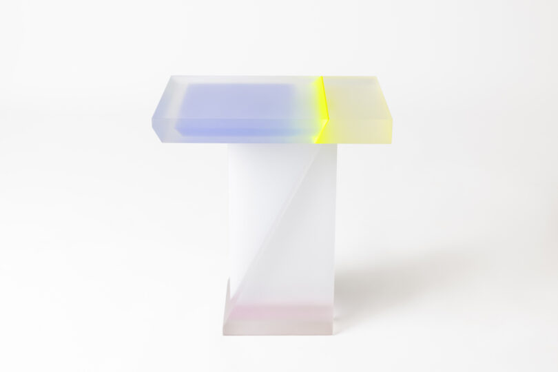 A translucent modern table with a rectangular tabletop that has a gradient from blue to yellow, supported by a white base.