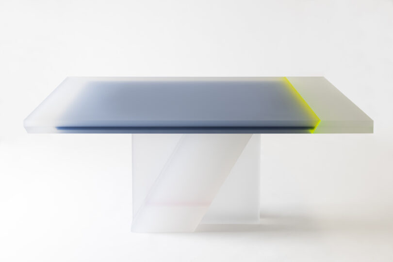 A modern rectangular table with a thick, semi-transparent white base and a smooth surface. The tabletop features a subtle gradient with hints of green and gray.