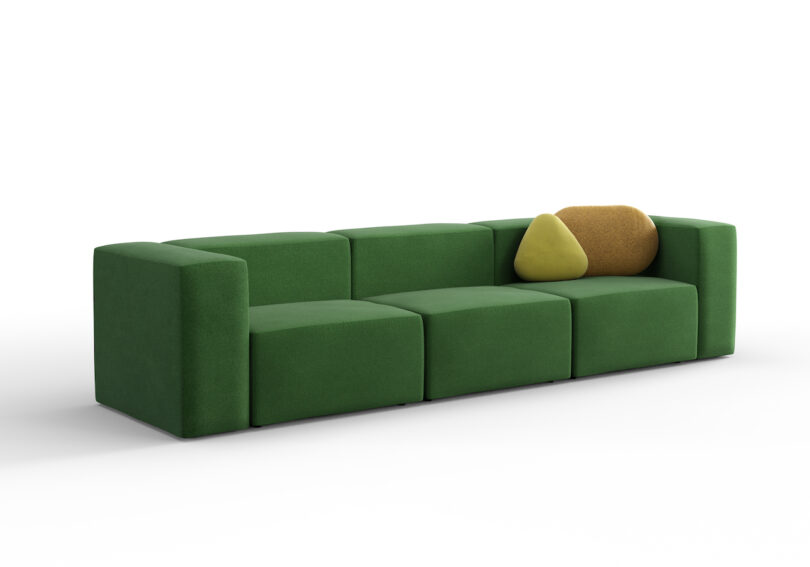 A modern green modular sofa with clean lines, featuring a yellow and a brown throw pillow. The sofa has a minimalist design with a low back and block-style cushions