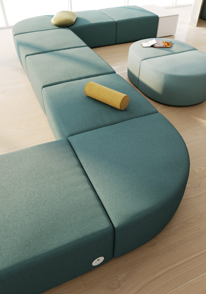 A modern modular seating arrangement with teal upholstered benches and a circular ottoman on a light wood floor. A yellow bolster pillow and a magazine on the ottoman are visible
