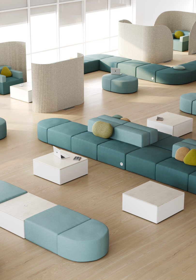A modern lounge area with teal modular seating, white tables, and a few yellow and green cushions, all arranged in a spacious, well-lit room with wooden floors