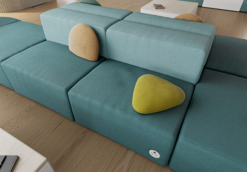 Modular teal seating units with mustard and beige triangular cushions arranged on top, set on a wood floor beside a white side table