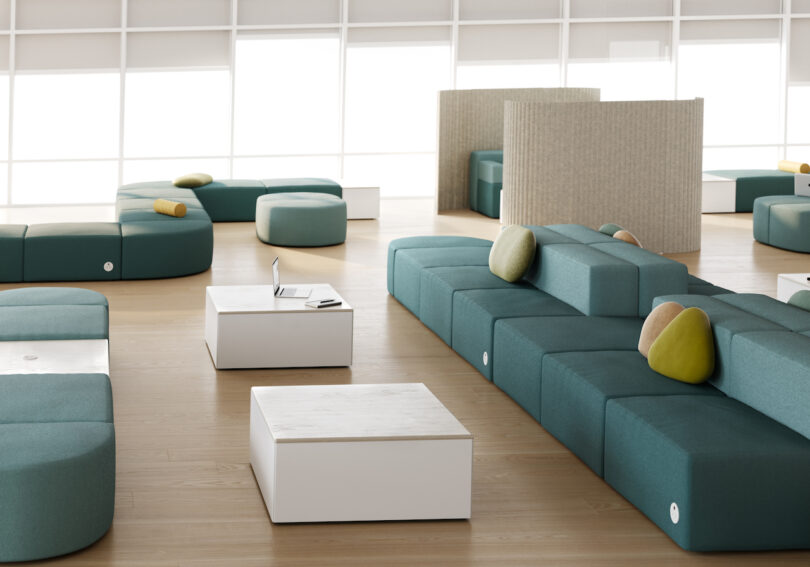 A modern office lounge features teal modular seating, white tables, and a laptop. Natural light streams through large windows and partitions provide semi-private areas
