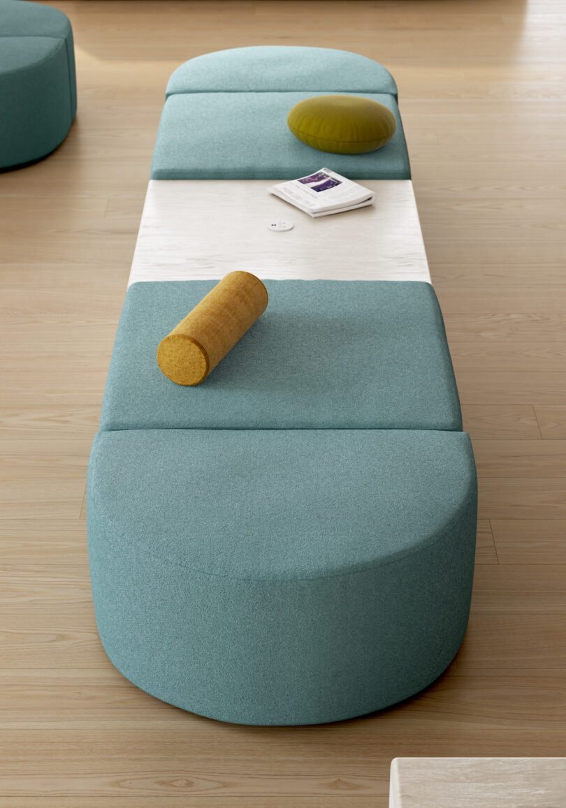 A modern bench with teal cushions and two pillows, one round yellow and one cylindrical yellow. A small table in the middle holds a book and a round white device. The floor is light wooden