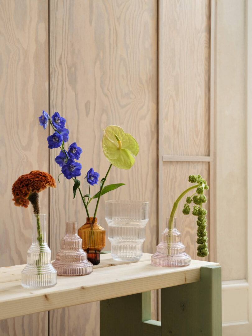 A wooden table with various glass vases, each containing different types of flowers, including blue delphiniums, a green anthurium, and others, set against a wooden textured background