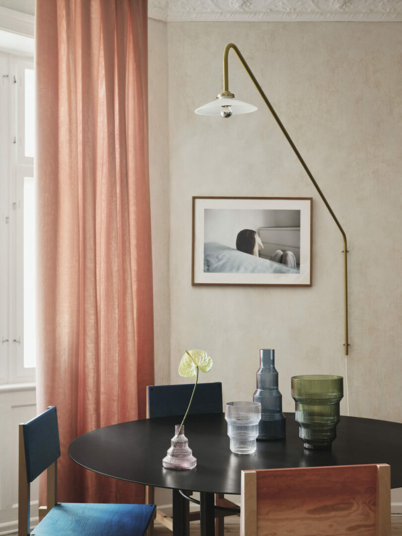 A dining area with a round black table, assorted glass vases, a single leafy plant, surrounding chairs, a framed photo on the wall, and a minimalist wall-mounted light fixture. Pink curtains adorn the window