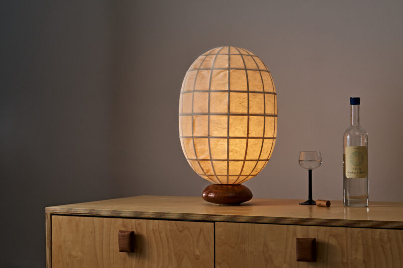 A lit, white, oval-shaped lamp on a wooden stand is placed on a wooden dresser. To the right, there's a bottle of clear liquid, a small glass, and a cork. The background is a plain wall