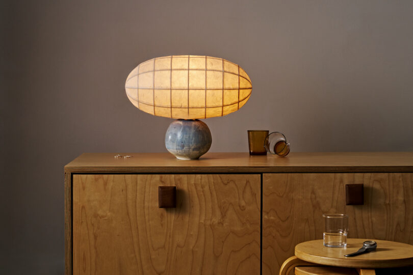 A wooden cabinet with leather handles displays a paper lamp turned on, ceramic vase, and two tumbler glasses. A round wooden stool with a glass and spoon is placed next to it, all set against a plain wall