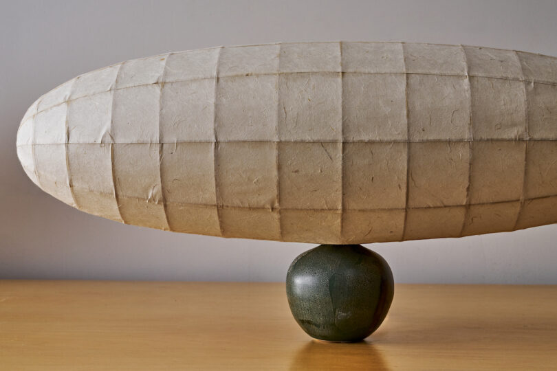 A rounded paper lamp is mounted on top of a small green ceramic base, placed on a wooden surface.
