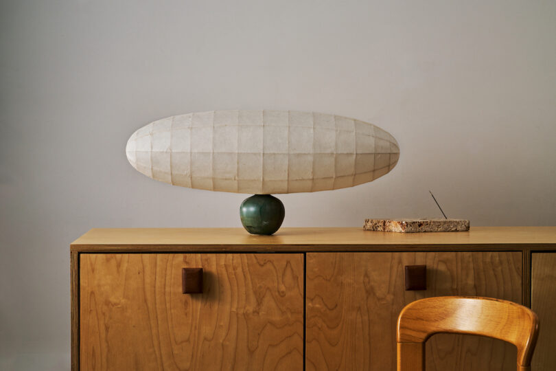 A large, oval-shaped lamp sits atop a green base on a wooden cabinet. An incense holder with incense is placed beside it. A wooden chair is partially visible in the foreground