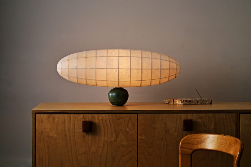 A lit, large, oval-shaped lamp sits atop a green base on a wooden cabinet. An incense holder with incense is placed beside it. A wooden chair is partially visible in the foreground.